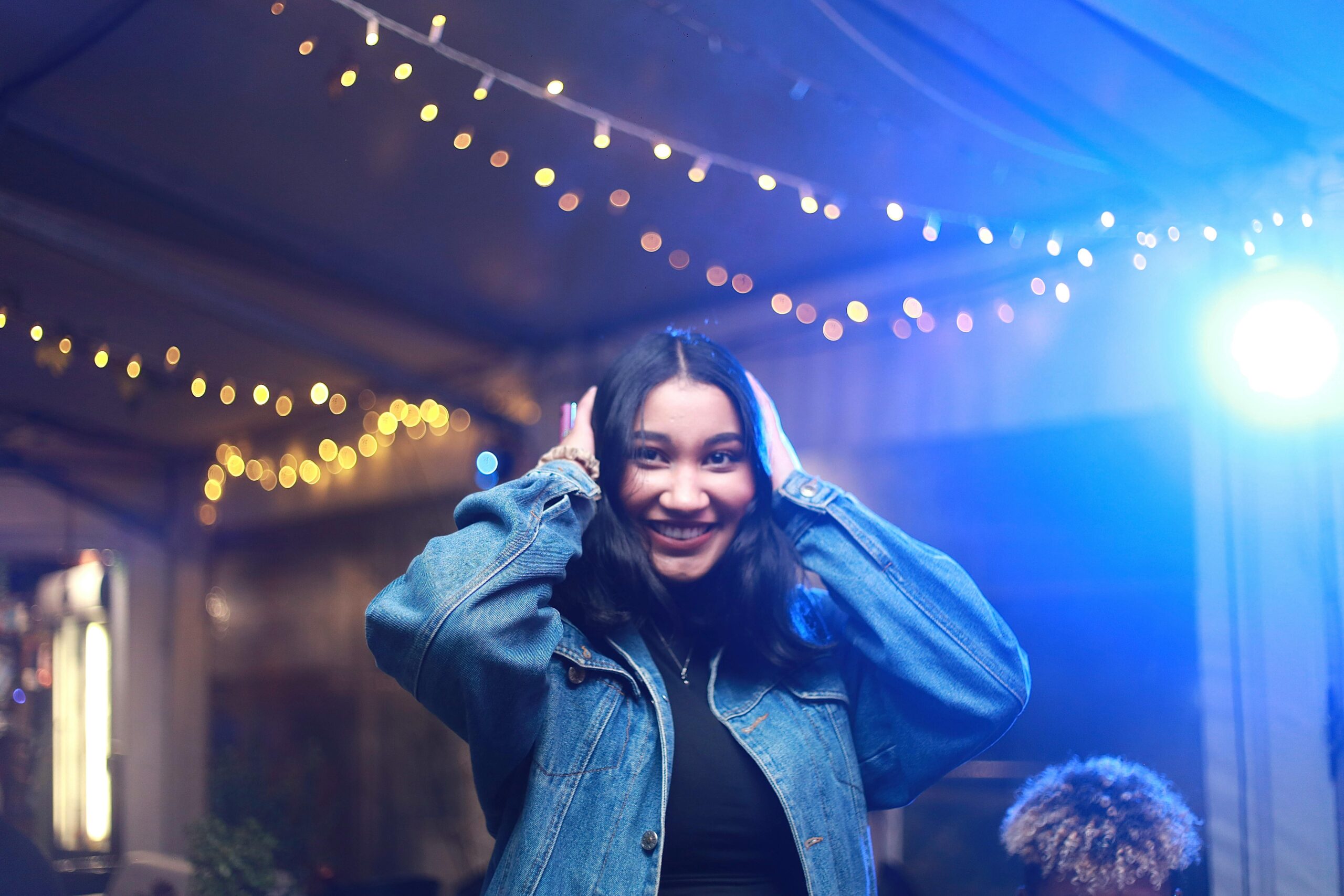 Woman with black hair wearing a jean jacket clutching her head while smiling.