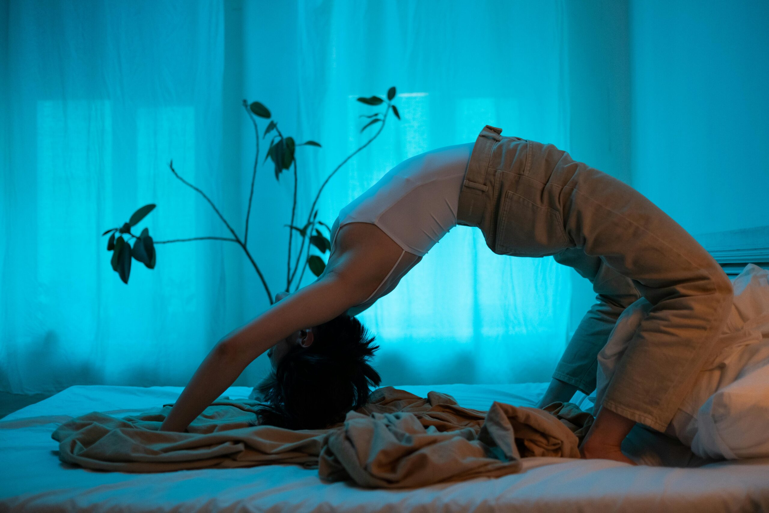 woman wearing a white tank top and khaki pants doing a backbend on a bed with a plant in the background