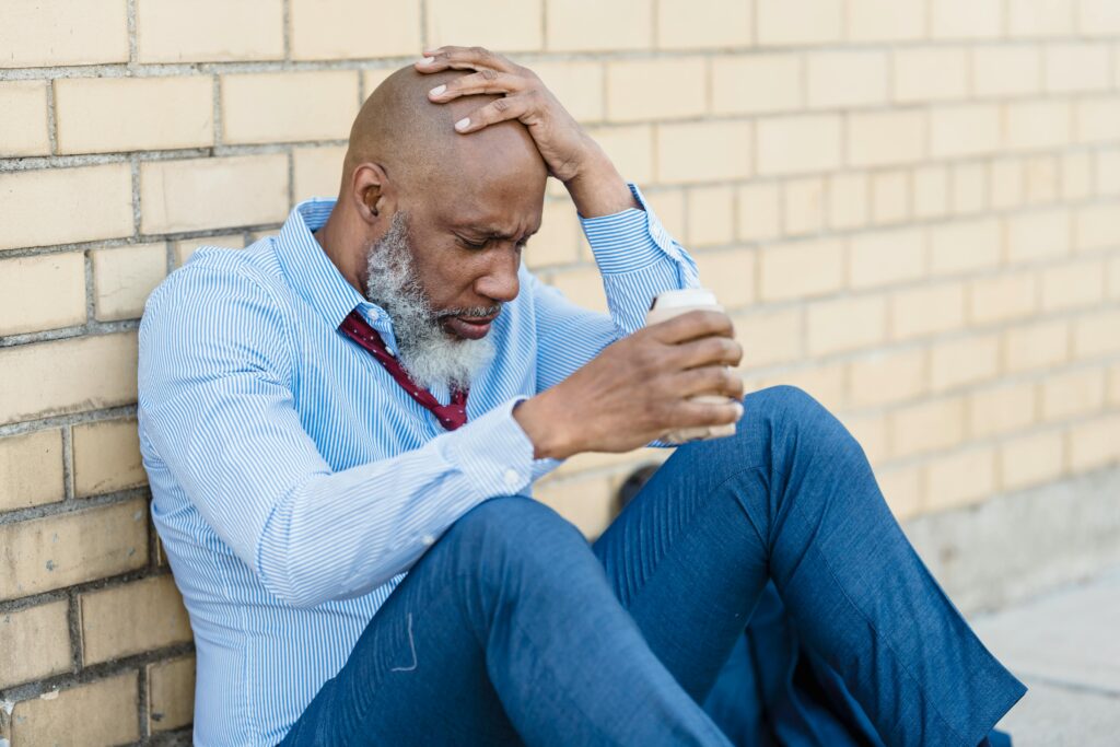 middle aged Man sitting on the ground with a drink in his hand, wearing a blue button up shirt and jeans. He has his hand on his head. What Is Brain Fog After Drinking? North Star Treatment