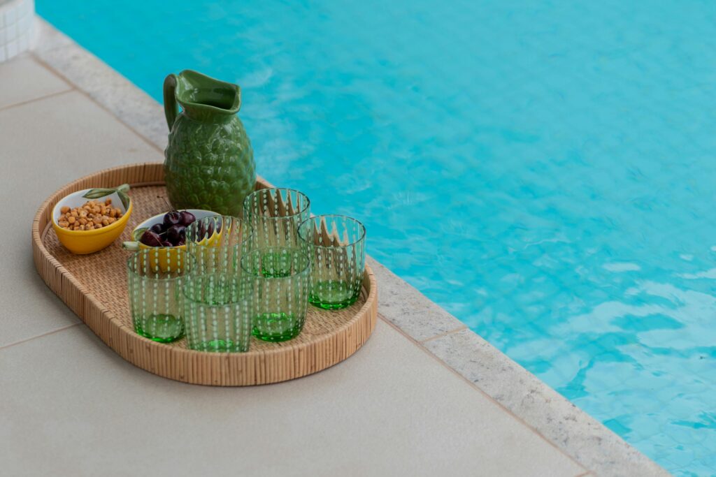 Image of a pool with a stand that has green glass cups and a green pitcher as well as finger food snacks. Luxurious living.