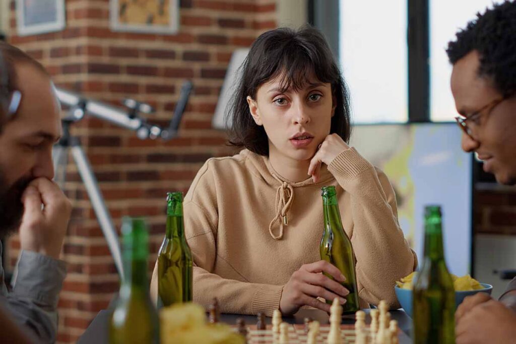 A woman sits at a table holding a beer bottle, looking concerned, surrounded by peers—symbolizing the early signs of substance misuse addressed at Northstar Treatment Center.